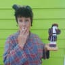 “A person in a plaid robe and patterned headscarf holds a cigarette to their mouth while displaying a small handmade doll dressed similarly and also holding a cigarette, against a bright green wooden wall.”