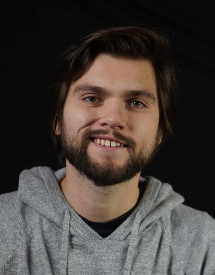 A man with medium-length brown hair and a short beard smiles at the camera. He wears a gray hoodie and is photographed against a plain black background.