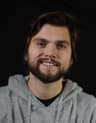 A man with medium-length brown hair and a short beard smiles at the camera. He wears a gray hoodie and is photographed against a plain black background.
