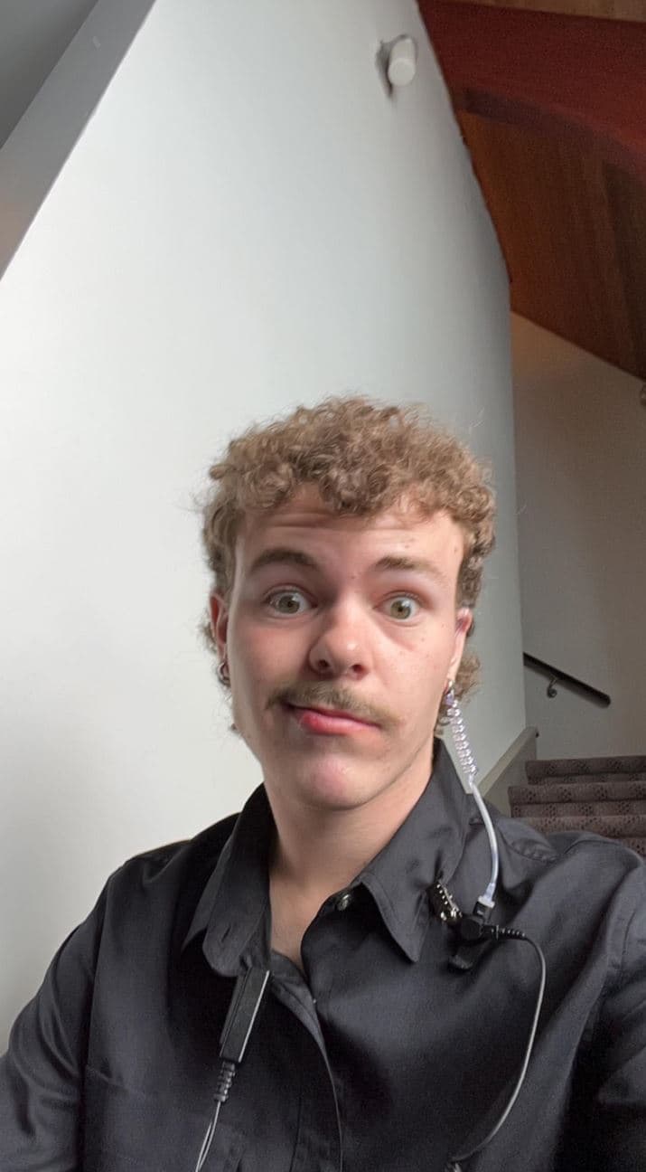 A person with short curly hair and a mustache looks at the camera with a raised eyebrow expression. They wear a black button-up shirt and an earpiece with a coiled wire. A staircase and light-colored wall are visible in the background.