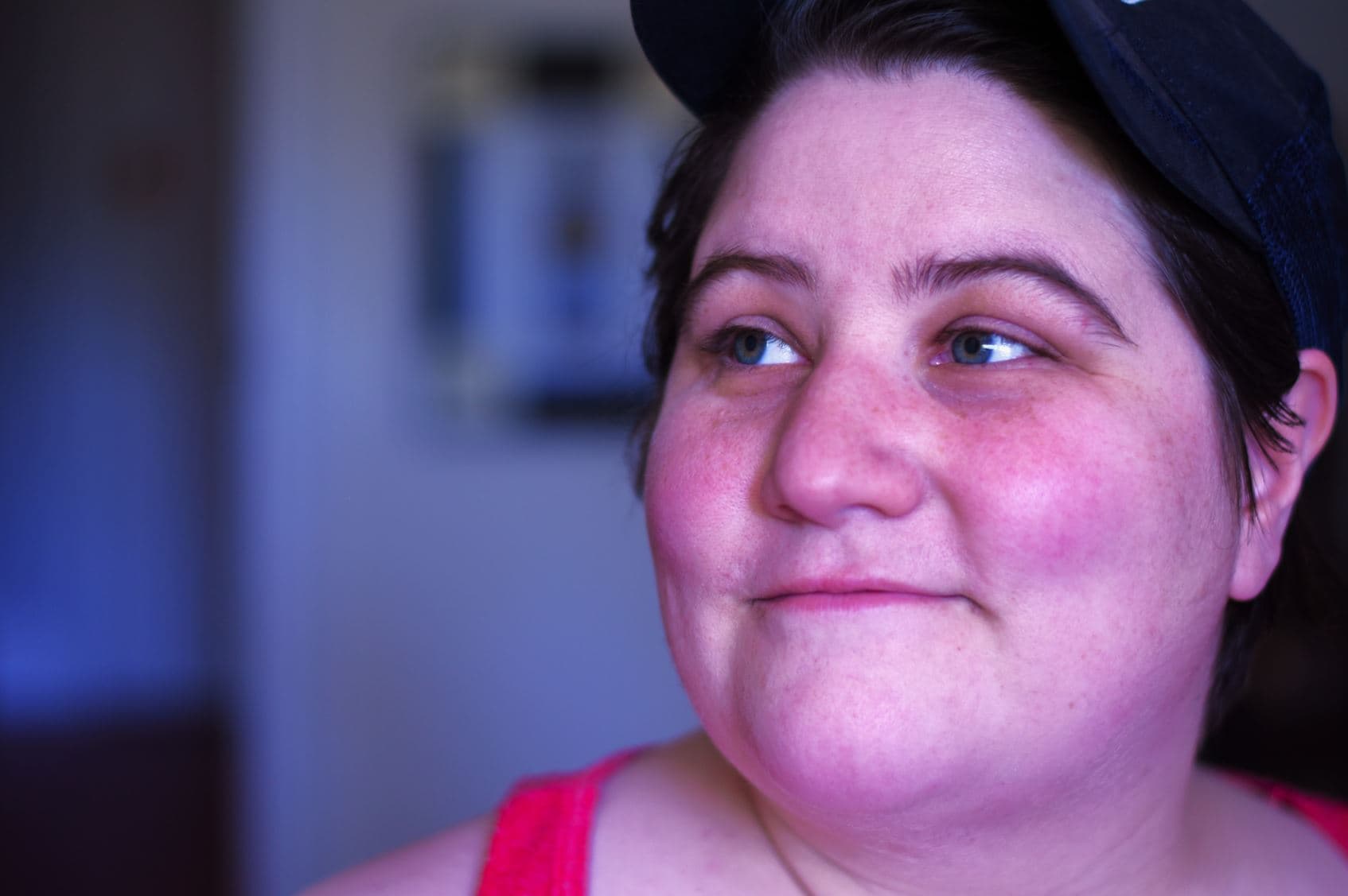 Close-up of a person wearing a black cap and red top, looking slightly to the side. They have short dark hair, light-colored eyes, and a neutral expression. The background is softly blurred, suggesting an indoor setting.