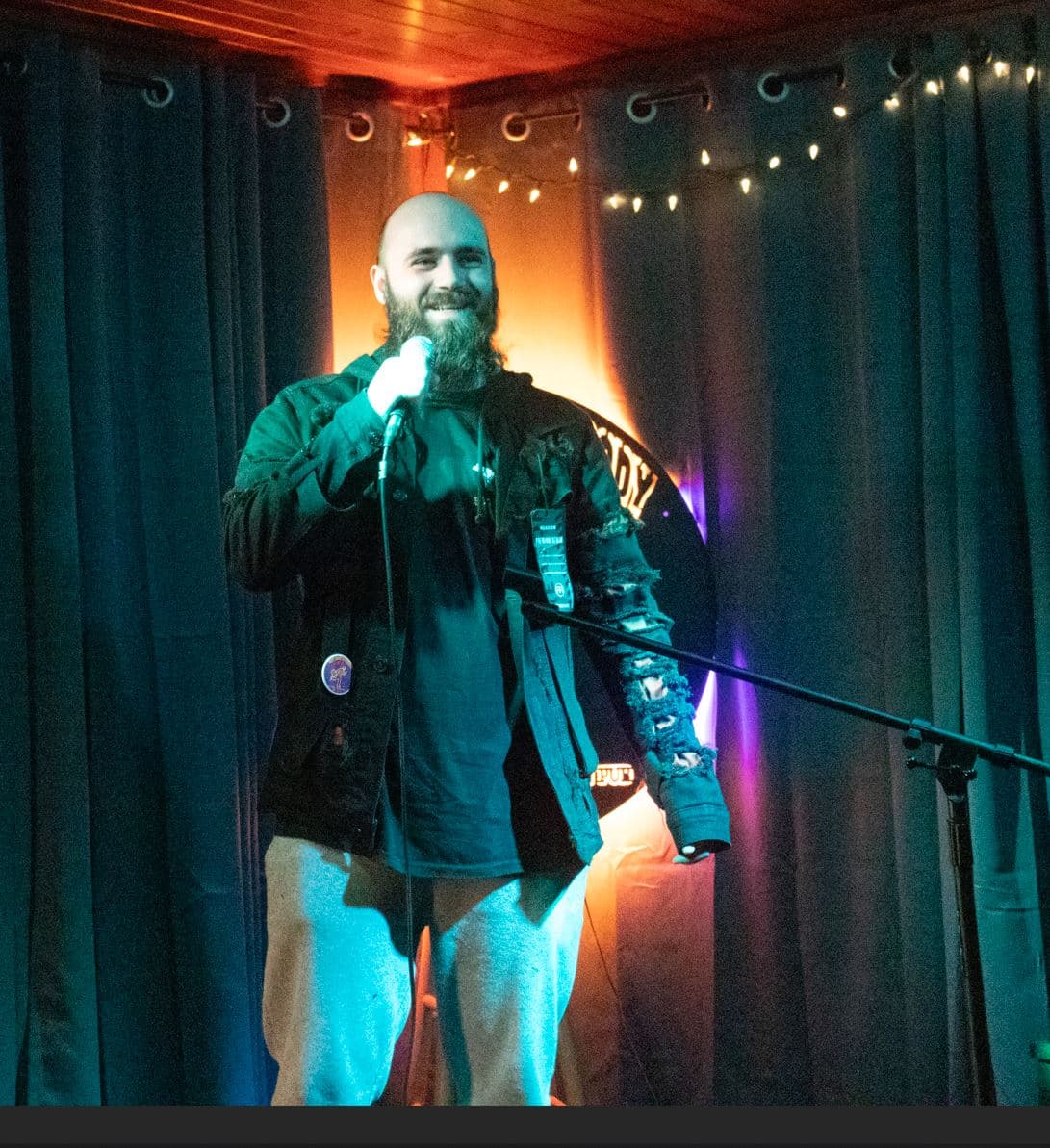 A person with a shaved head and beard stands on a small stage holding a microphone. They wear a dark shirt, light pants, and a distressed jacket. Colored lights and string lights hang in front of dark curtains behind them.
