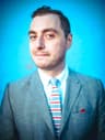 A man in a gray suit jacket and white shirt poses against a bright blue background. He wears a striped tie with red, white, and blue bands and a red pocket square. He looks slightly to the side with a neutral expression.