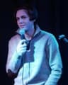 A person stands on stage holding a microphone, wearing a light-colored sweater. Blue stage lighting casts across them, with a dark curtain and a mic stand visible in the background.