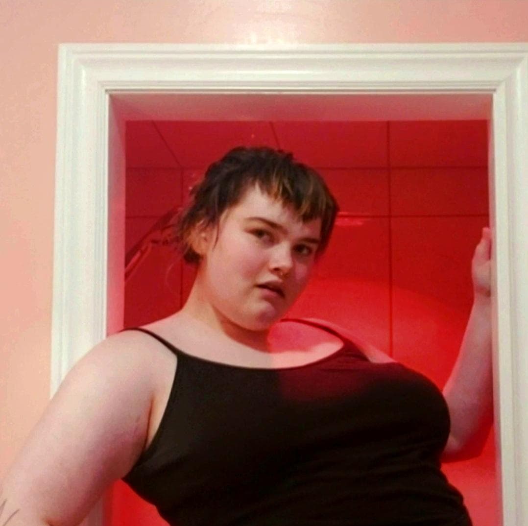 Person with short dark hair wearing a black tank top stands in a doorway lit with red lighting. One hand rests on the doorframe, and pink walls surround the doorway in the background.