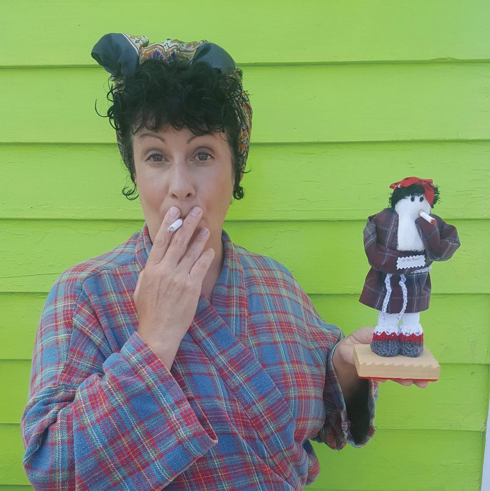“A person in a plaid robe and patterned headscarf holds a cigarette to their mouth while displaying a small handmade doll dressed similarly and also holding a cigarette, against a bright green wooden wall.”