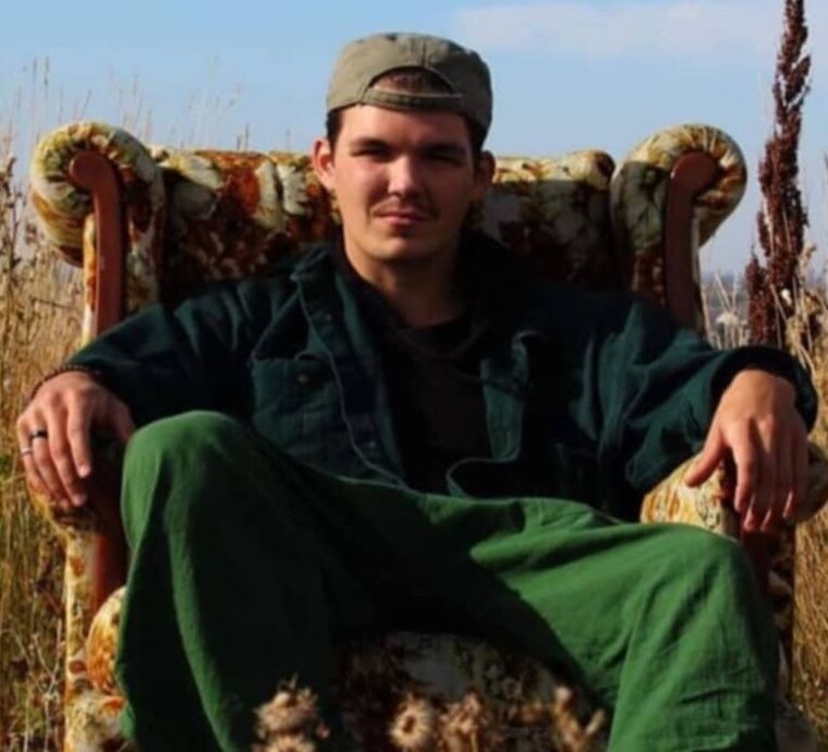 A person sits in a patterned armchair placed outdoors in a field of tall grass. They wear a backward cap, dark jacket, green pants, and sneakers, with hands resting on the chair arms and a neutral expression.