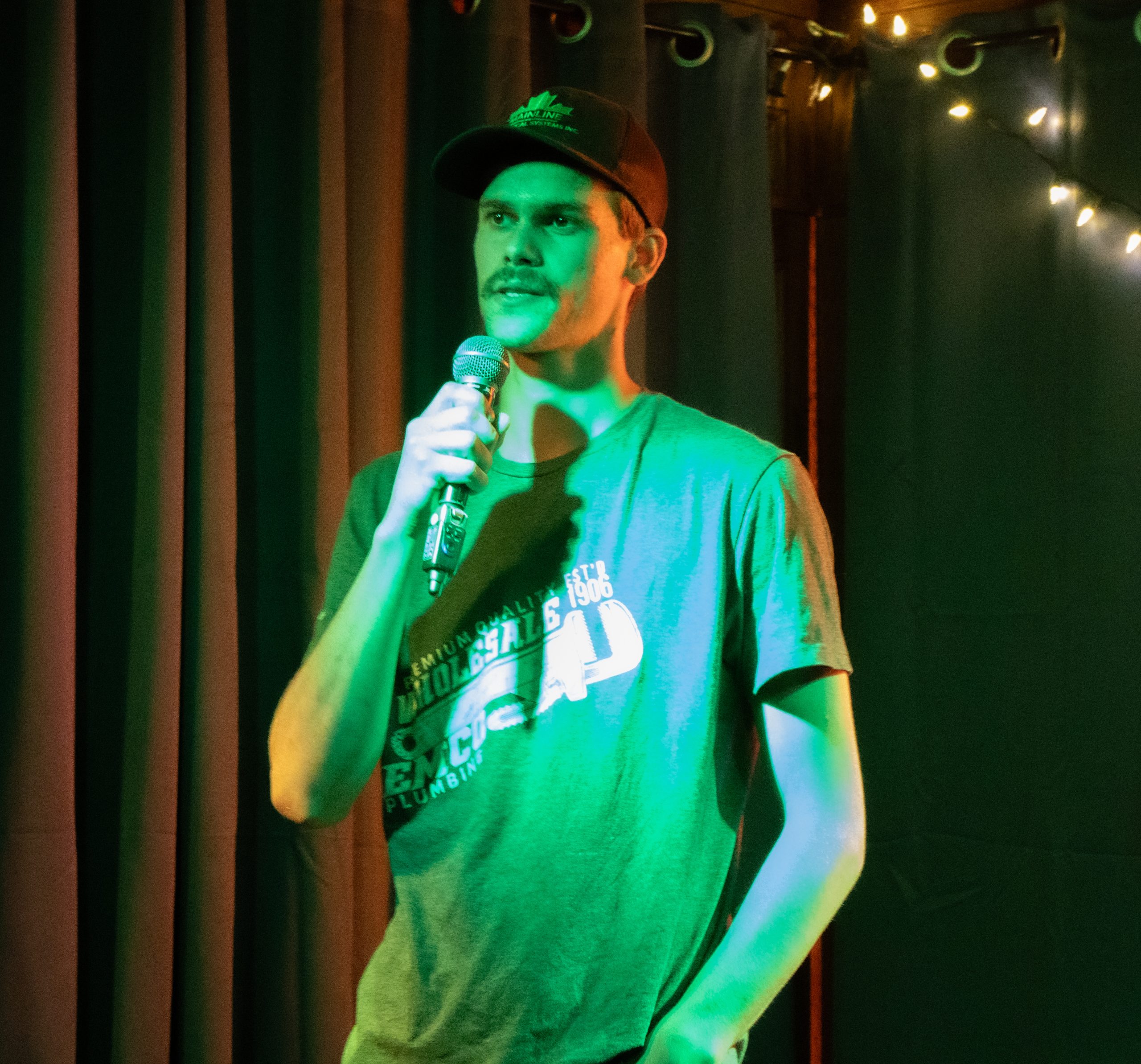 A person wearing a baseball cap and T-shirt stands on stage holding a microphone. Green stage lighting casts over them, with dark curtains and string lights visible in the background.