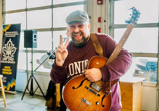 A man wearing a gray flat cap and maroon sweatshirt holds a hollow-body electric guitar and smiles while making a peace sign with his hand. He stands indoors near large garage-style windows, with a speaker and a sign visible in the background.